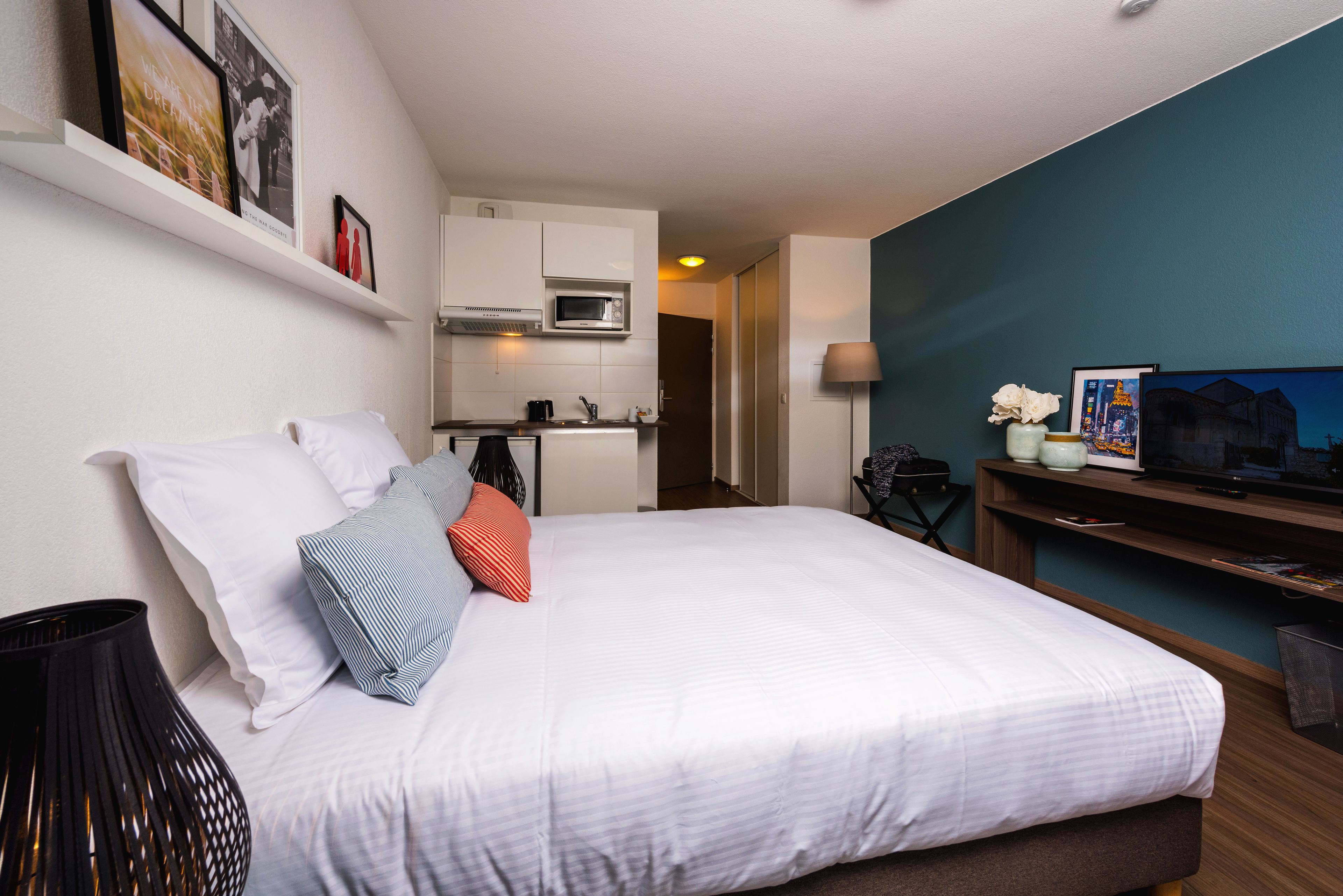 A bedroom with a large bed, a TV, a desk, and a kitchenette.