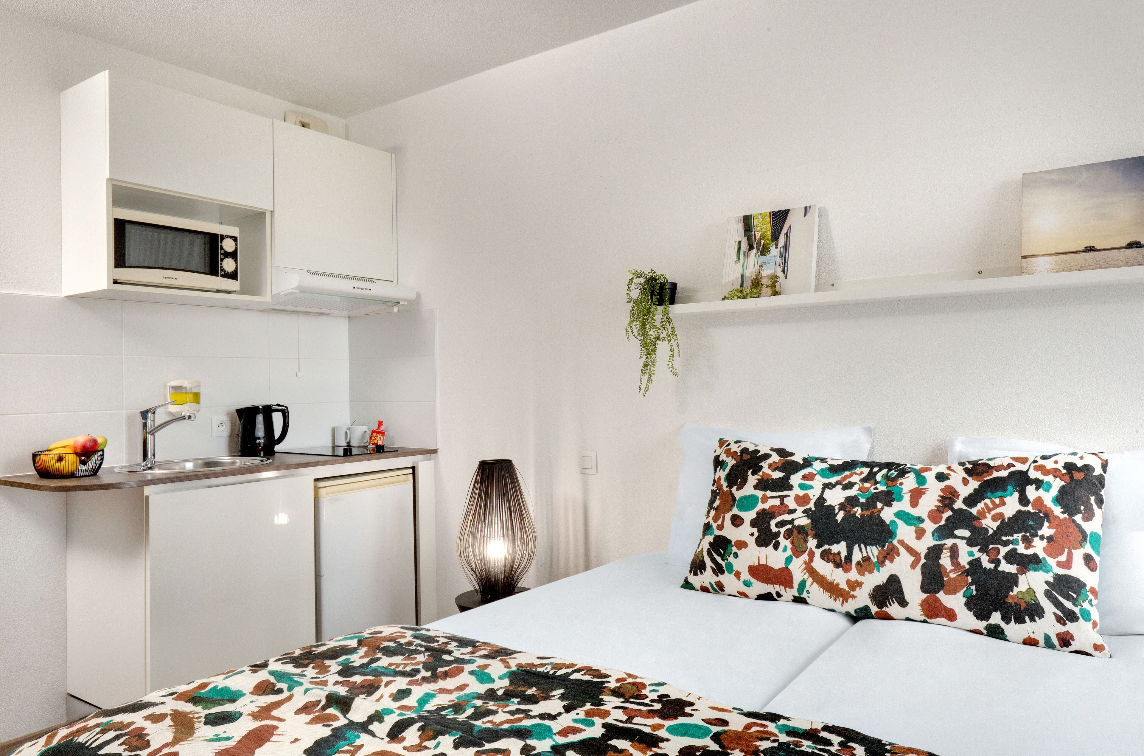 A compact hotel room with a kitchenette, a bed with patterned bedding, a bedside table with a lamp, and a wall-mounted shelf with decorative items.