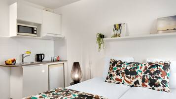 Habitación de hotel compacta con kitchenette, cama con ropa de cama estampada, mesita de noche con lámpara y estante en la pared con adornos.
