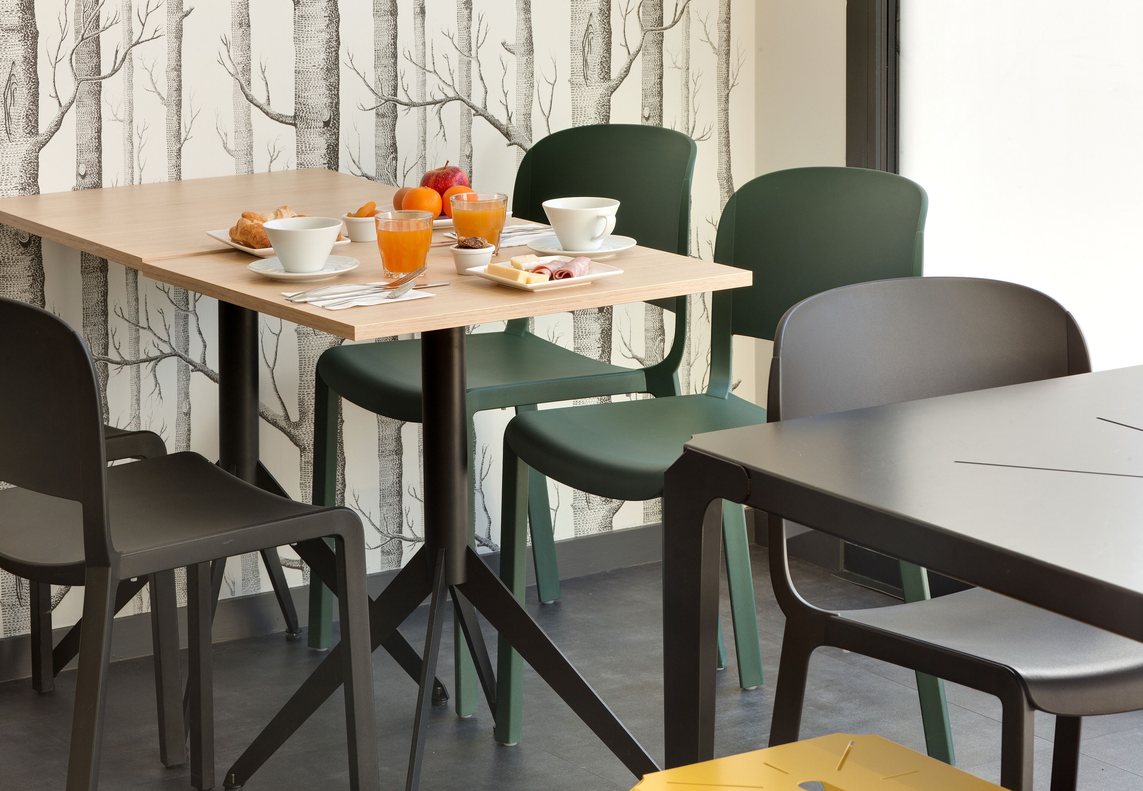 A breakfast table with croissants, fruit, and orange juice, surrounded by green and black chairs in a modern room with a tree-patterned wallpaper.