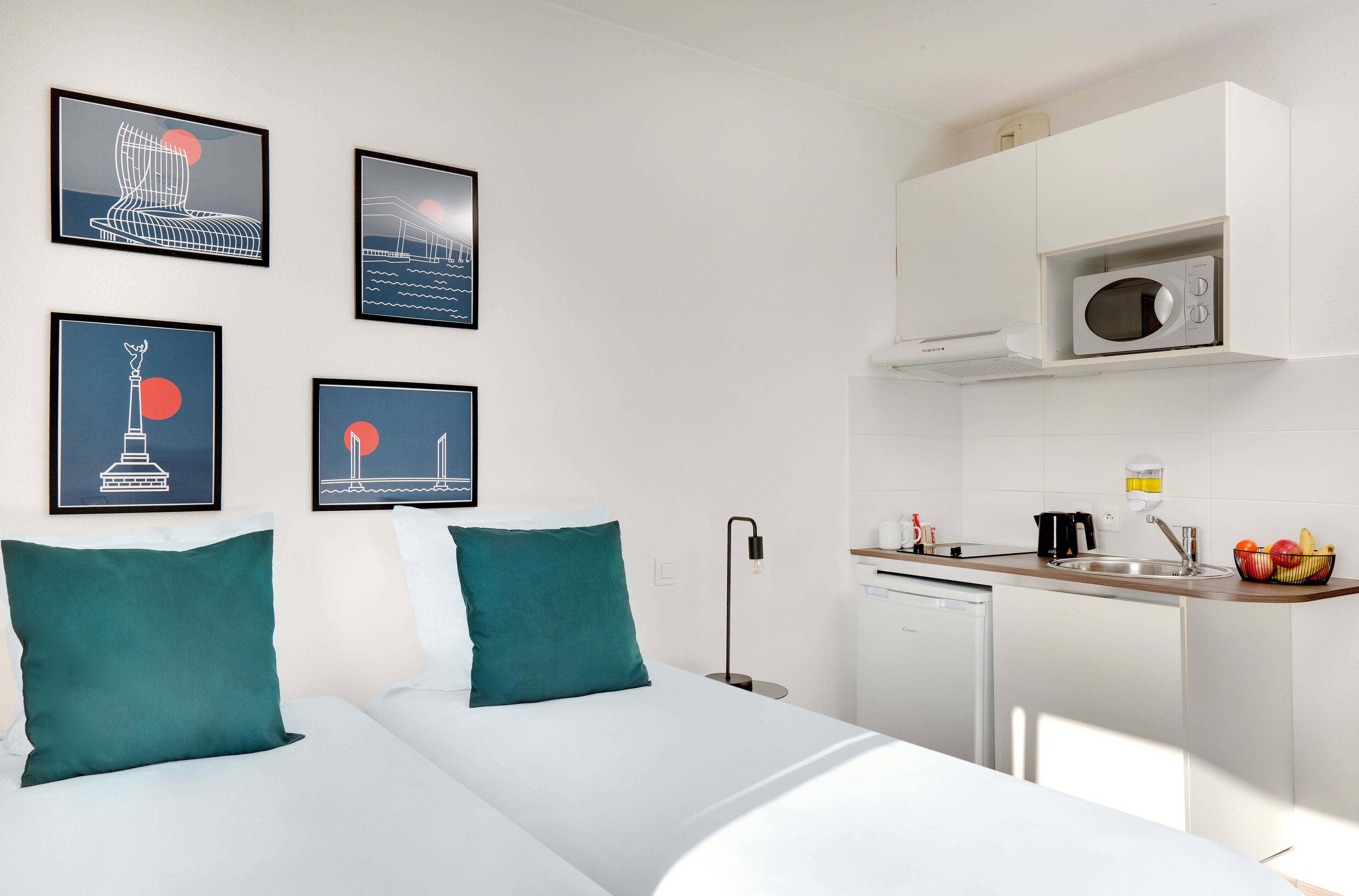 A modern hotel room with a bed, a small kitchenette, and artwork on the wall.