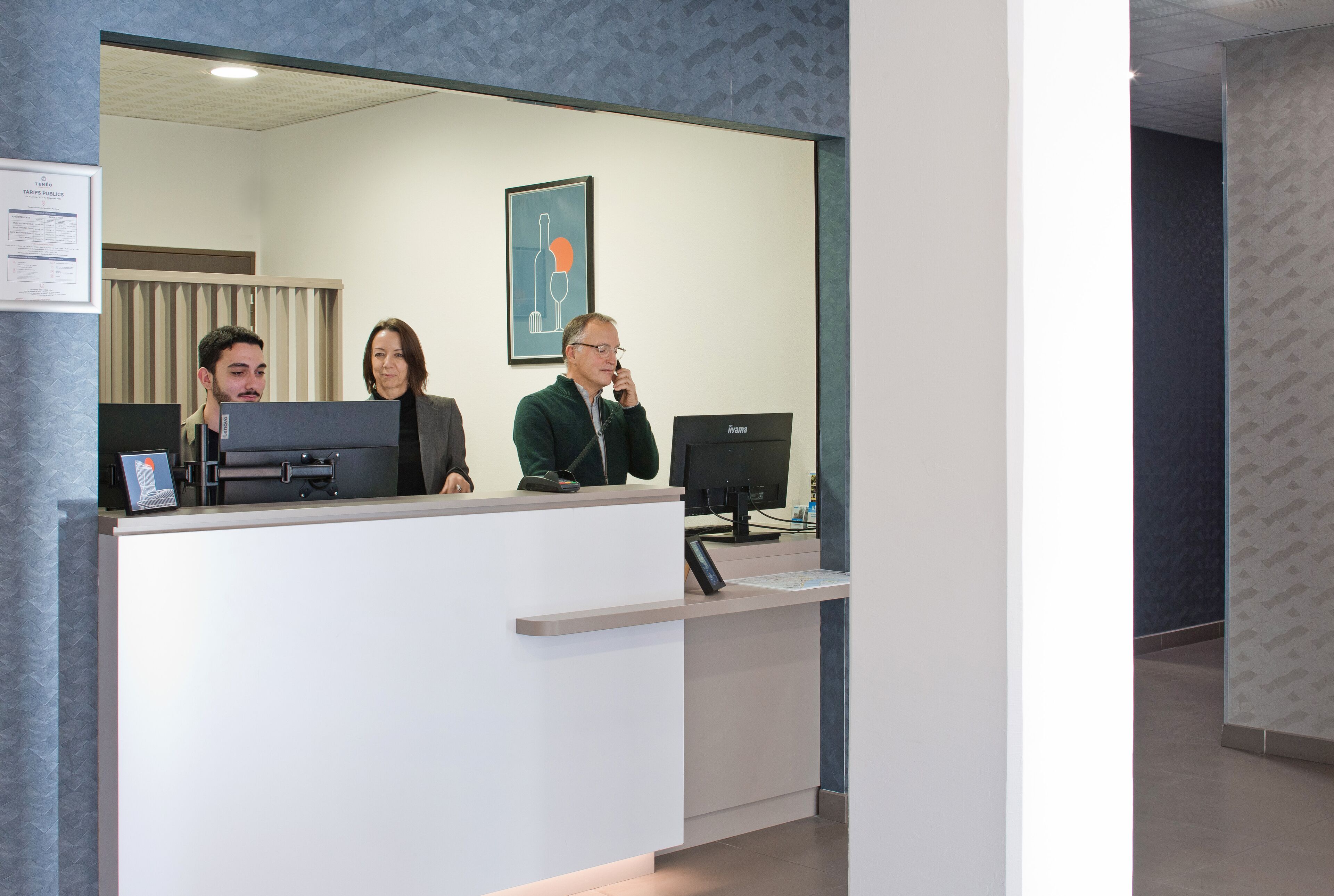 Una recepción con dos personas sentadas detrás, un certificado enmarcado en la pared y un monitor de computadora sobre el mostrador.