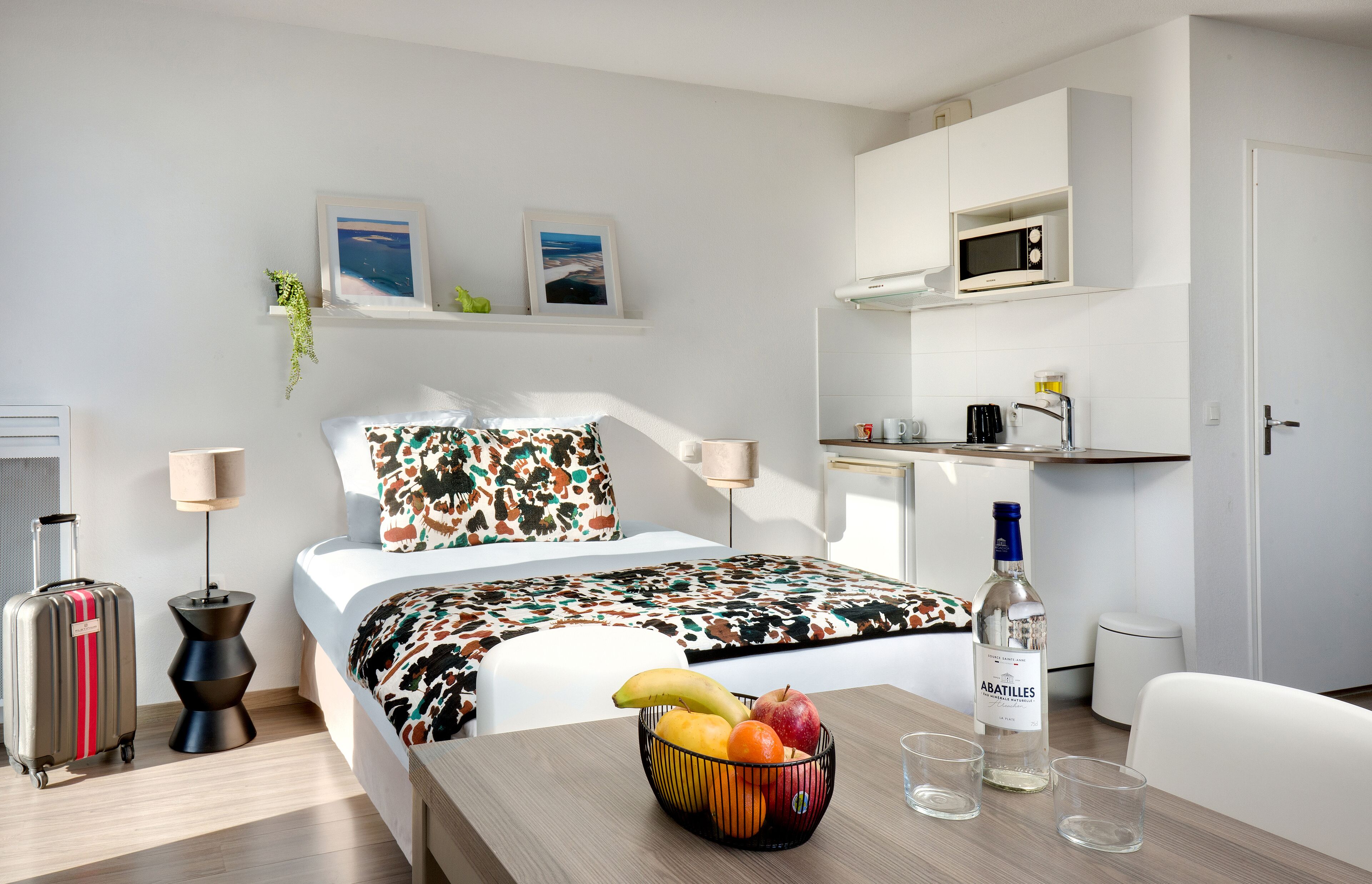 Une chambre d’hôtel avec un lit, une table de chevet avec des fruits et des verres, une valise, un four à micro-ondes et un petit coin cuisine.