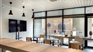 Un interior moderno con un largo banco de madera, mesas redondas y sillas, un televisor de pantalla plana montado en la pared y grandes ventanales que ofrecen vistas al exterior.