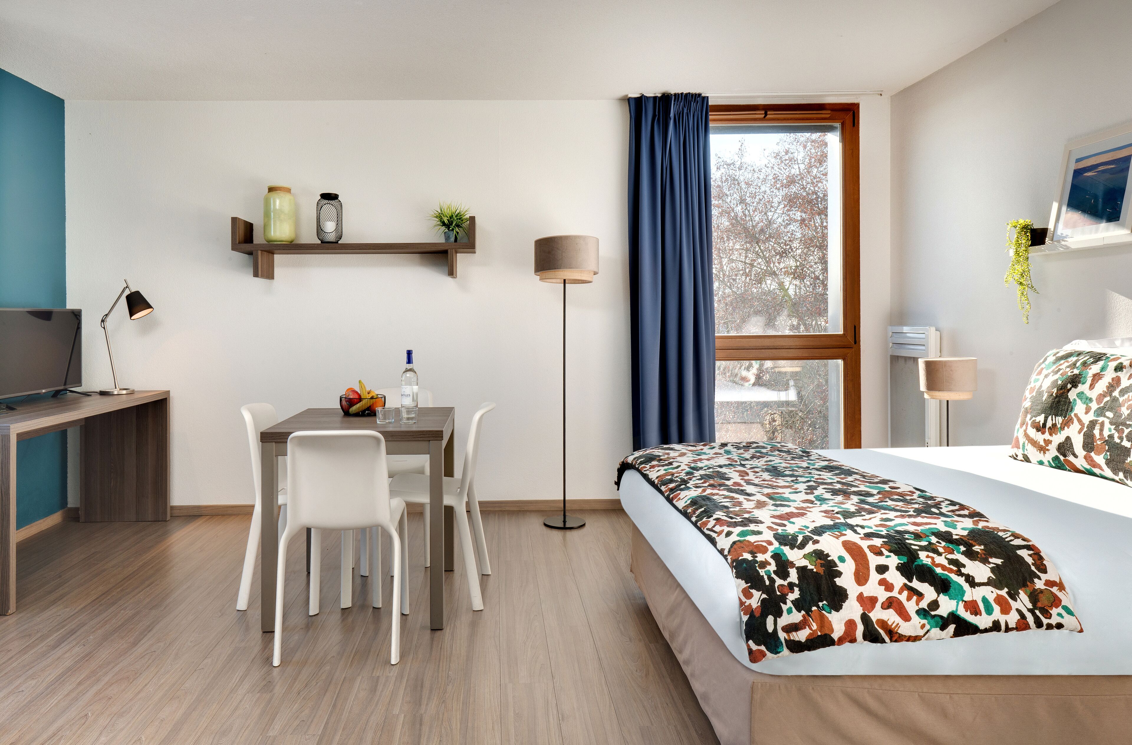 A modern hotel room with a wooden desk, white chairs, a bed with a colorful blanket, a standing lamp, and a window with a view of trees.