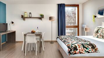 A modern hotel room with a wooden desk, white chairs, a bed with a colorful blanket, a standing lamp, and a window with a view of trees.