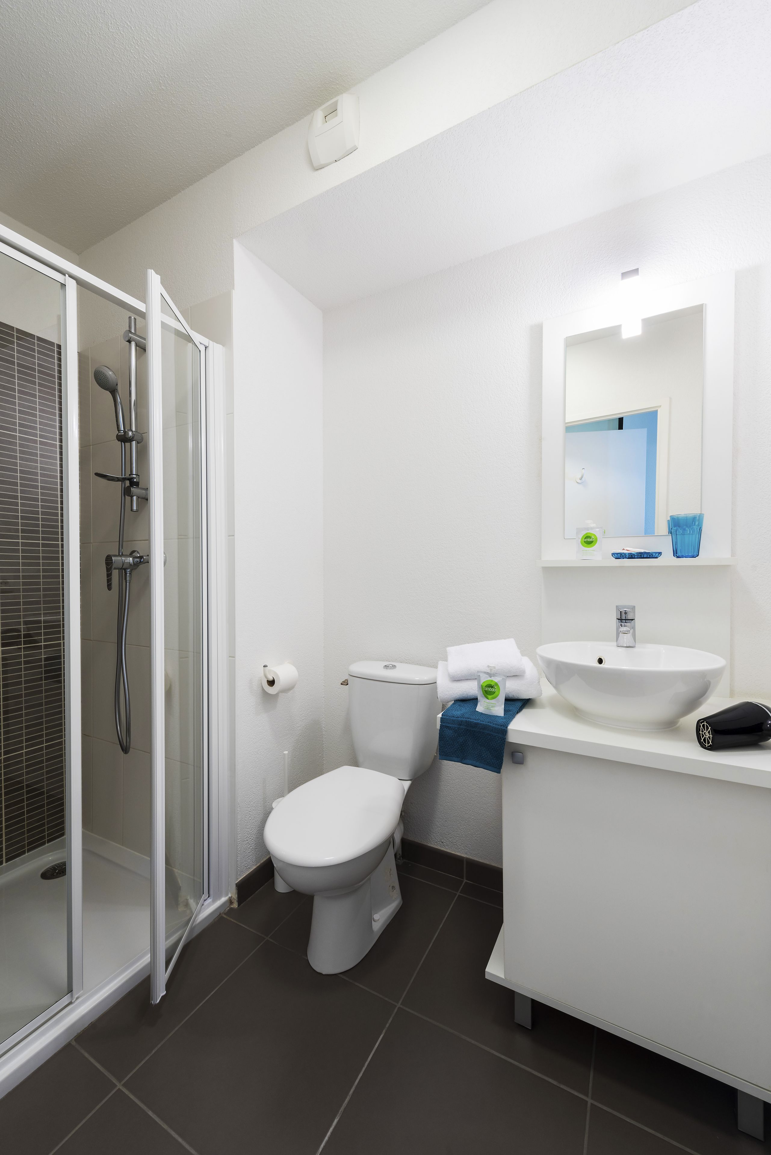 Un baño moderno con ducha envuelta en vidrio, inodoro blanco, lavamanos con espejo y una toalla azul sobre el mostrador.