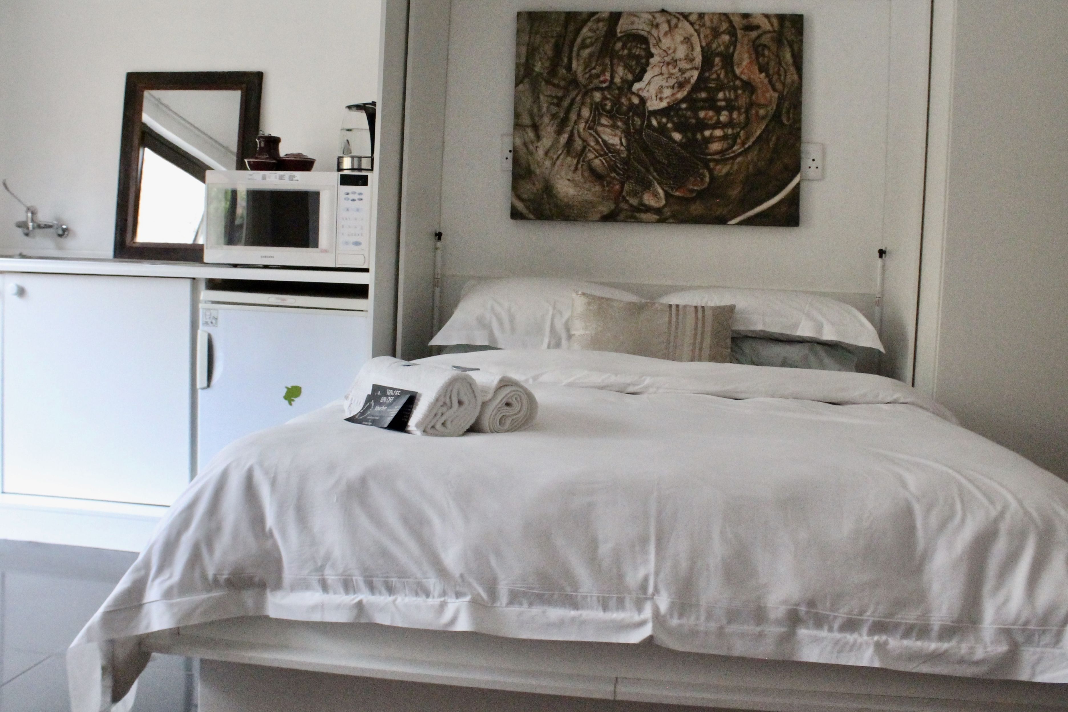 Un lit bien fait, avec du linge de lit blanc, une petite table de chevet sur laquelle se trouvent un livre et une bouteille, et une œuvre d’art accrochée au mur au-dessus du lit.