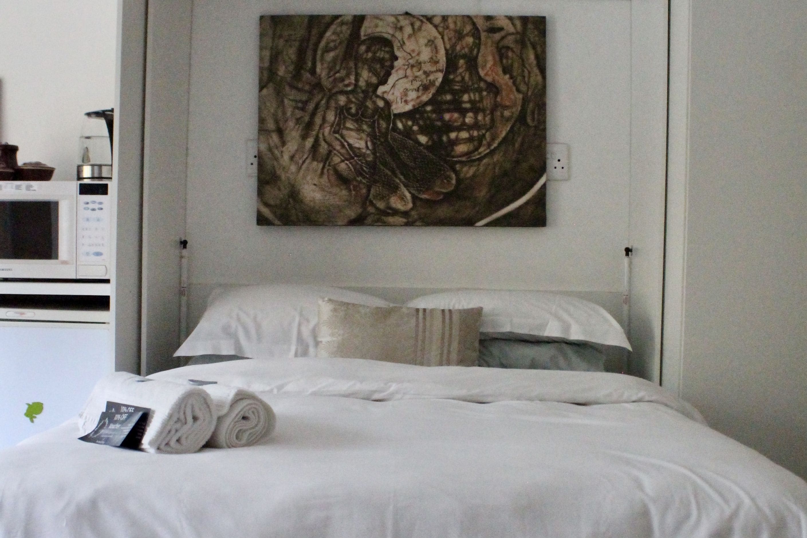 A neatly made bed with white linens and a rolled towel, a wall-mounted artwork depicting a skull, and a microwave on the left.
