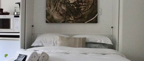 A neatly made bed with white linens and a rolled towel, a wall-mounted artwork depicting a skull, and a microwave on the left.