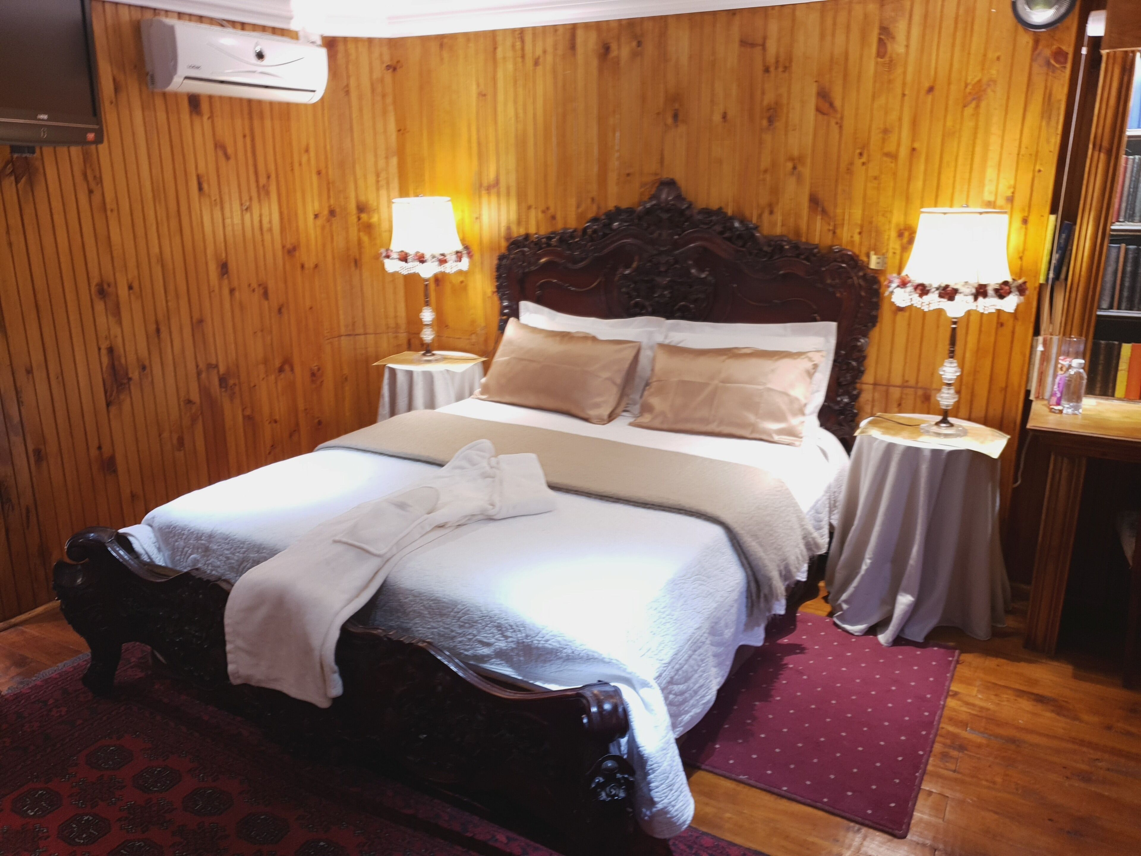 A wooden-paneled room with a large bed, two bedside tables with lamps, and a bookshelf.