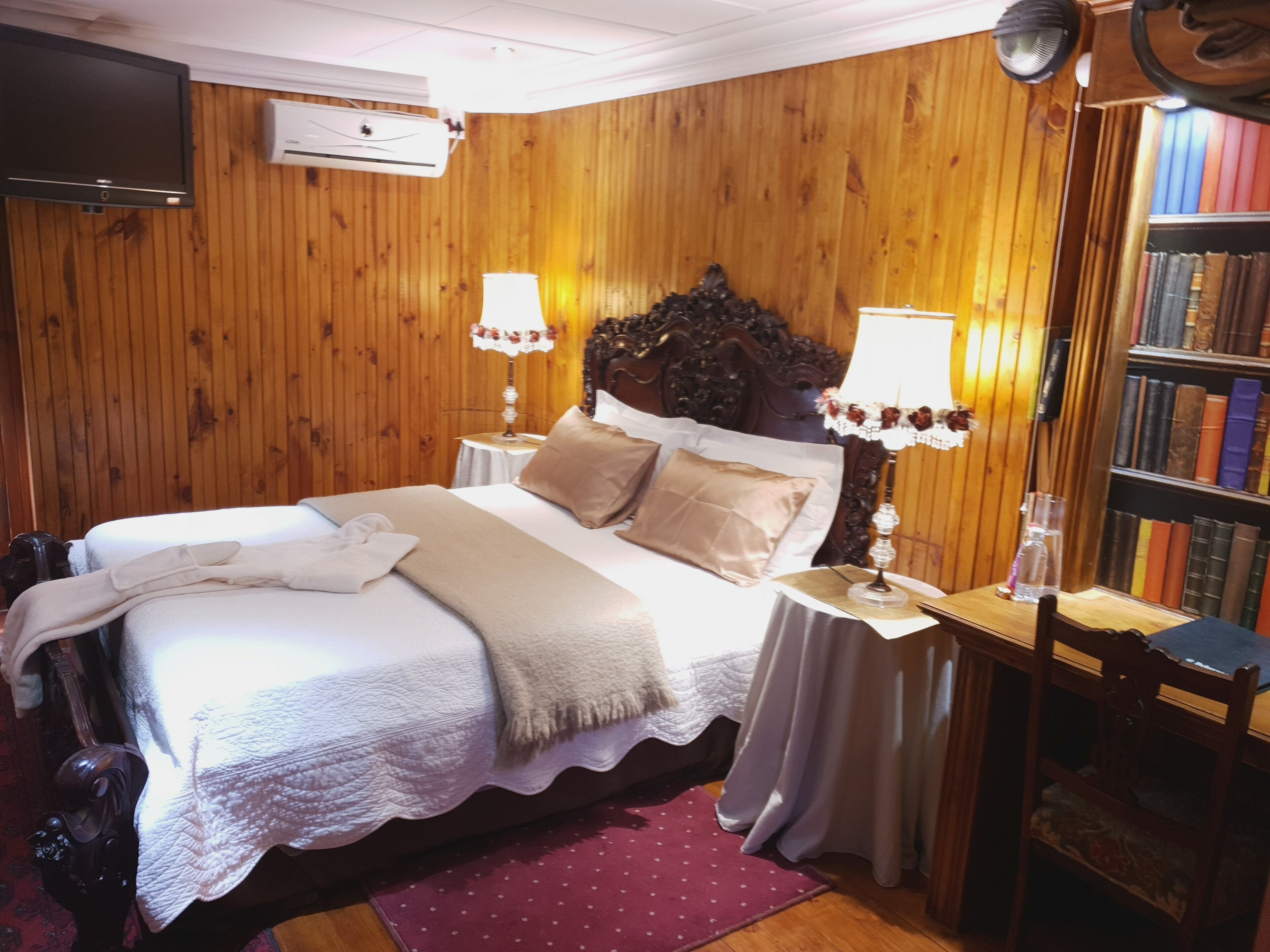 A hotel room with a wooden paneled wall, a large bed with a white comforter, two bedside tables with lamps, a bookshelf, and a flat-screen TV mounted on the wall.