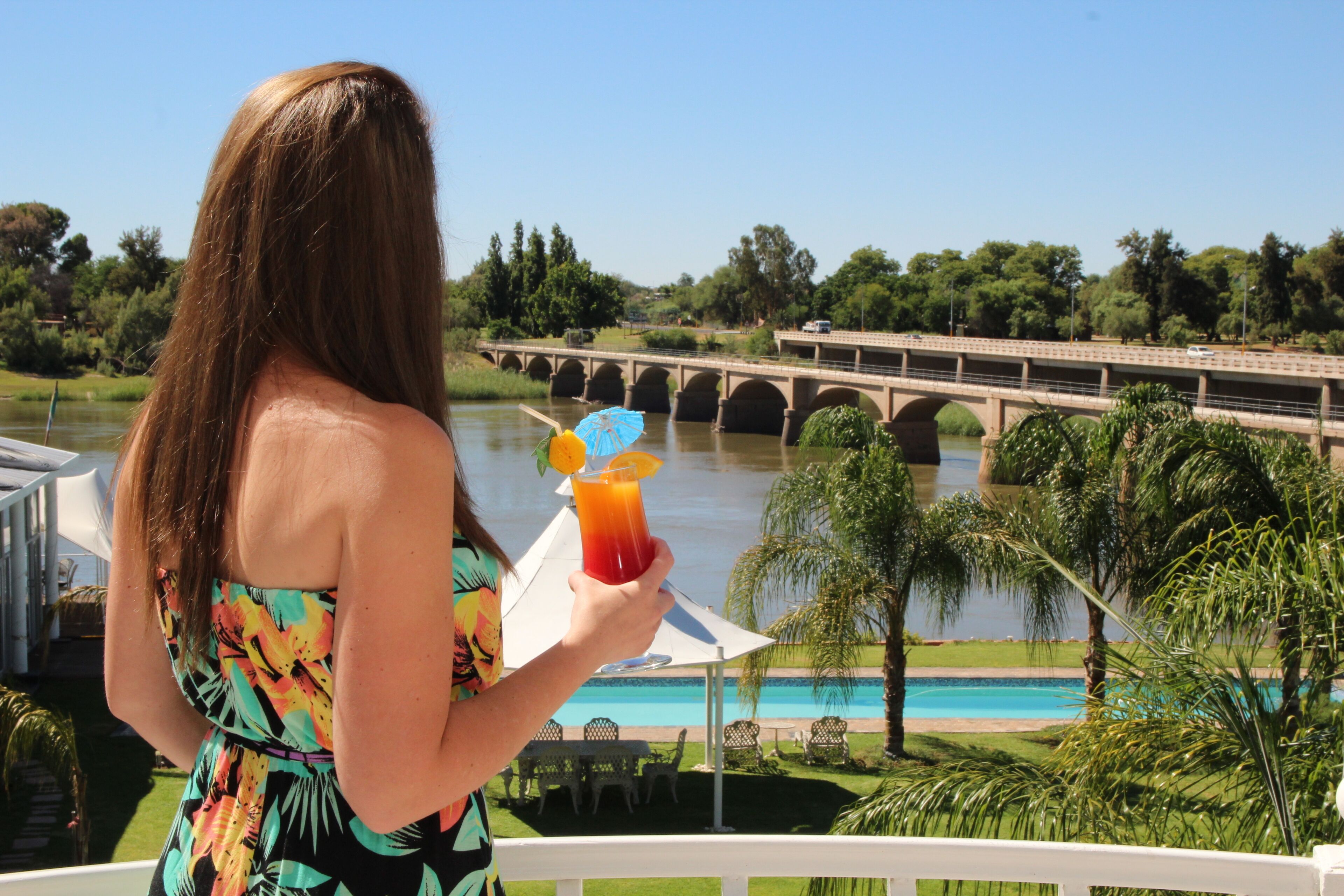 Una mujer sostiene una bebida con sombrilla tropical, con vistas a un puente y una piscina.