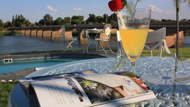 A glass of orange juice with a strawberry on the rim, a magazine on a glass table, and a view of a river with a bridge in the background.