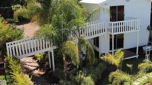 A two-story white house with a balcony, surrounded by lush greenery and palm trees.