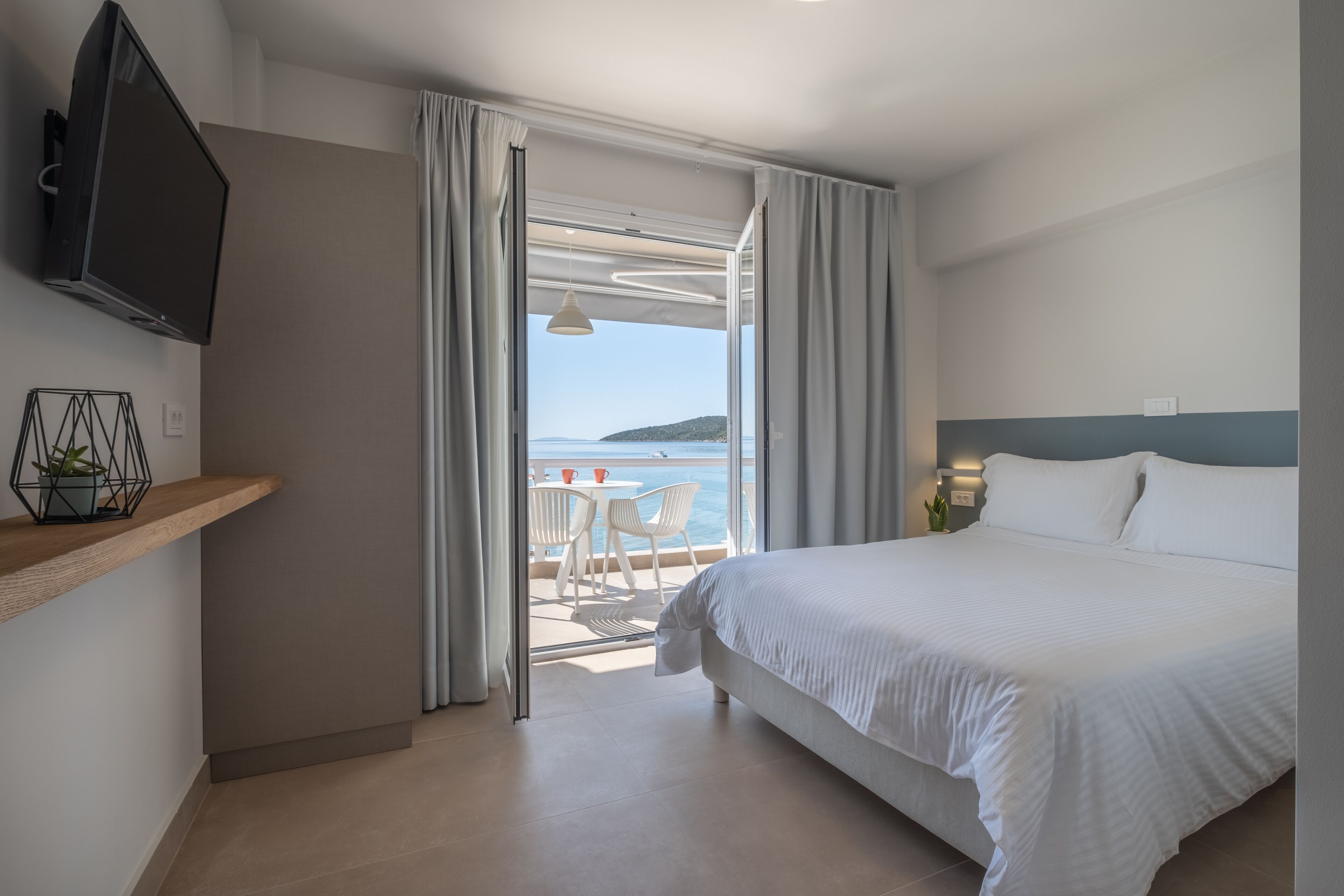 A hotel room with a bed, a TV, a balcony with a table and chairs, and a view of the sea.