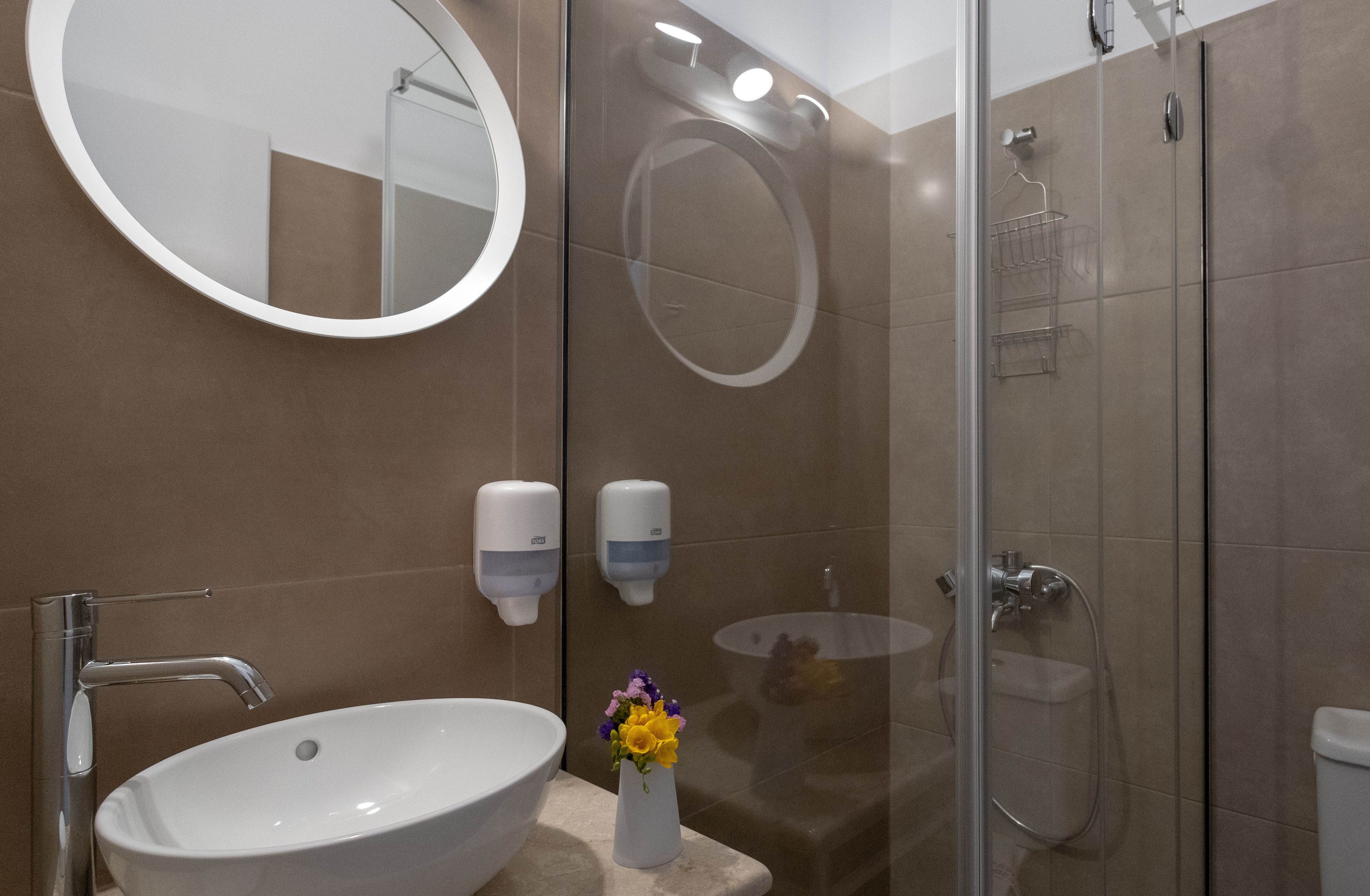 A modern bathroom with a round mirror, a sink, a glass-enclosed shower, and two soap dispensers.