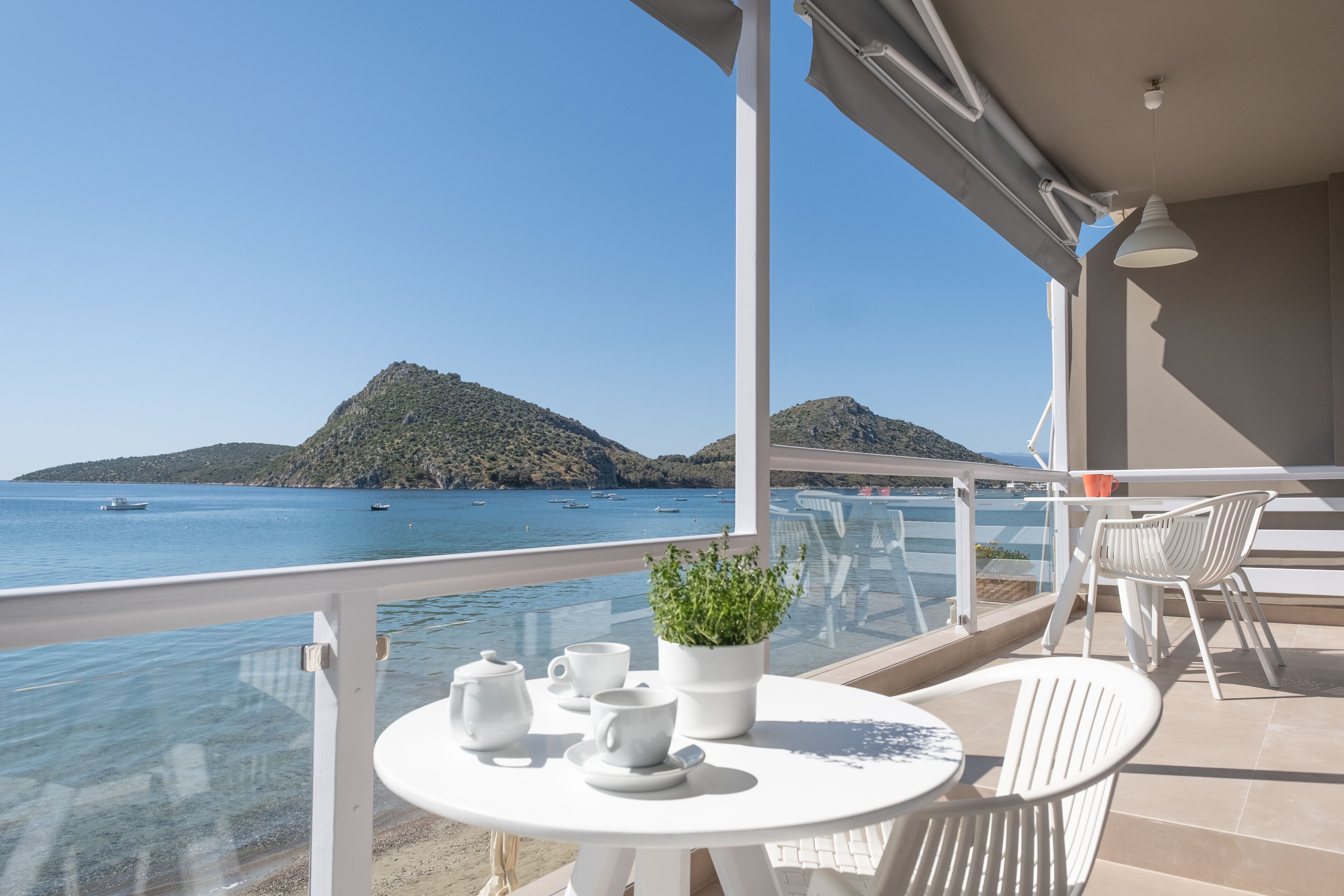 A balcony with a table set for tea, a potted plant, and a view of the sea and hills.