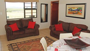 A living room with two brown sofas, a patterned rug, a table with a floral tablecloth, and a framed painting on the wall.