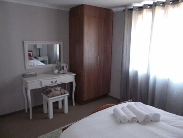 Quarto de hotel com guarda-roupa de madeira, uma secretária branca com espelho, um banquinho pequeno e uma cama com toalhas dobradas.