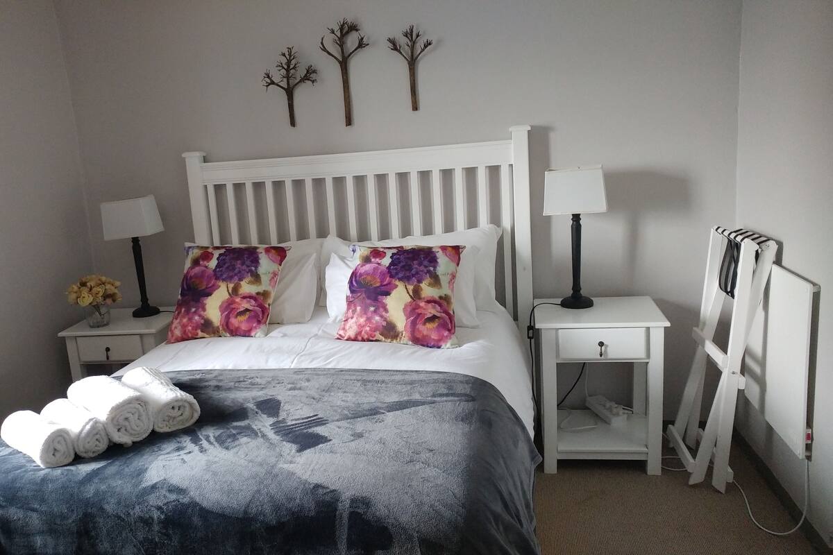 Een net opgemaakt bed met bloemenkussens, een nachtkastje met een lamp en een radiator.