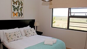 A neatly made bed with a teal blanket, white pillows with butterfly prints, and a view of a field through a window.