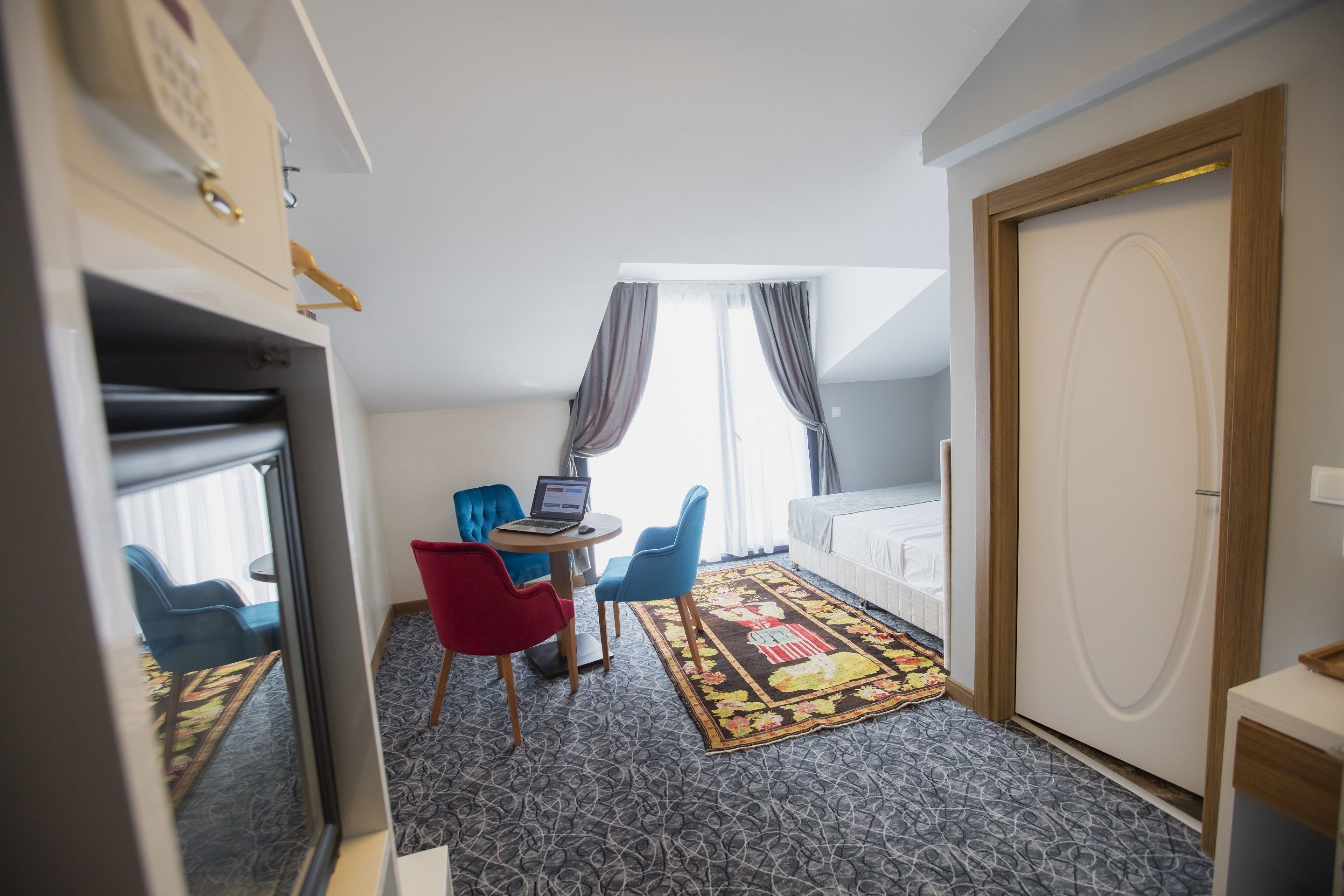 Une chambre d’hôtel avec un lit, un bureau avec un ordinateur portable, deux chaises, un petit tapis et une fenêtre avec des rideaux.