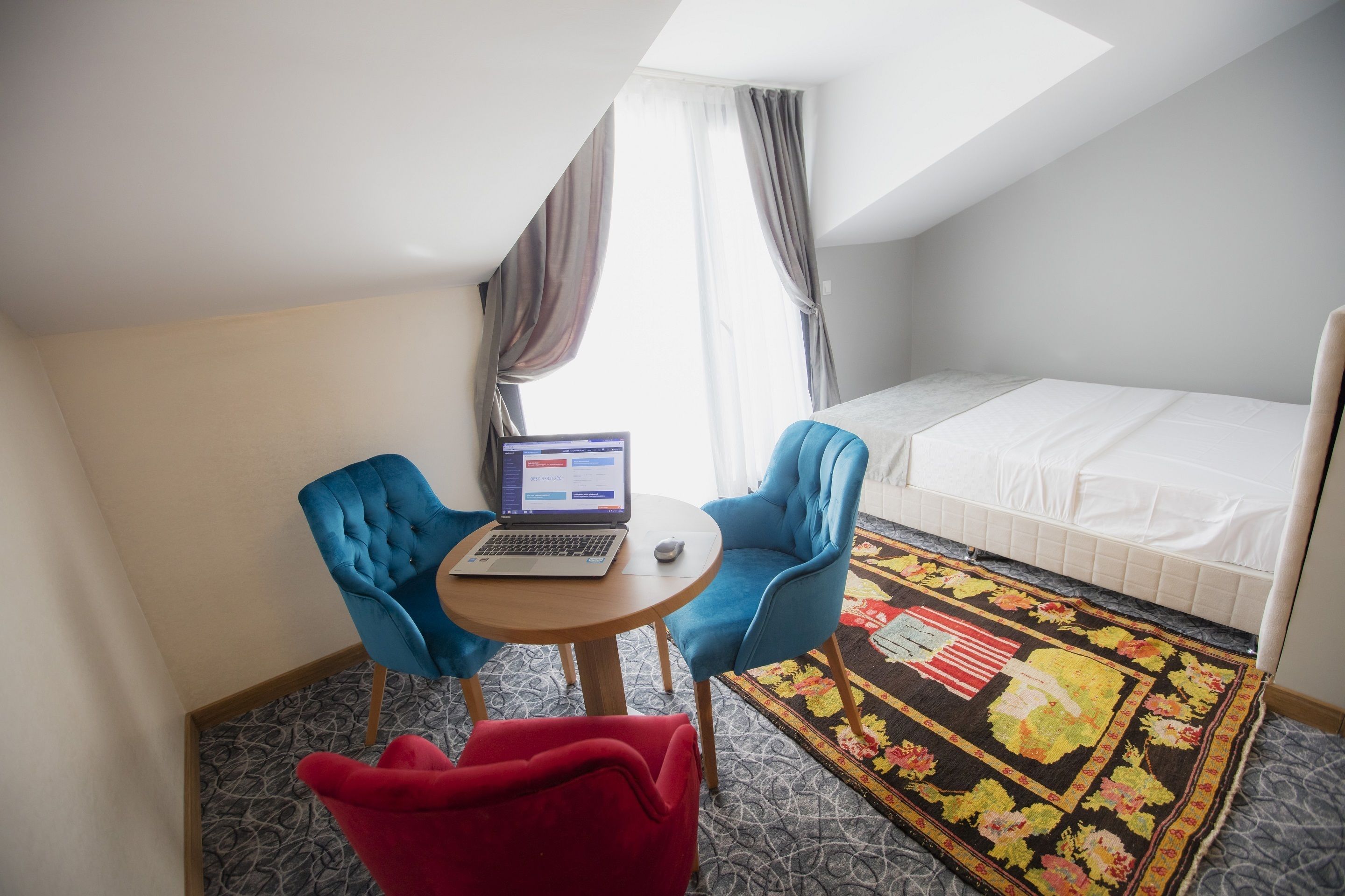 Een kleine, goed verlichte kamer met een bed, een bureau met een laptop, twee stoelen en een kleurrijk vloerkleed.