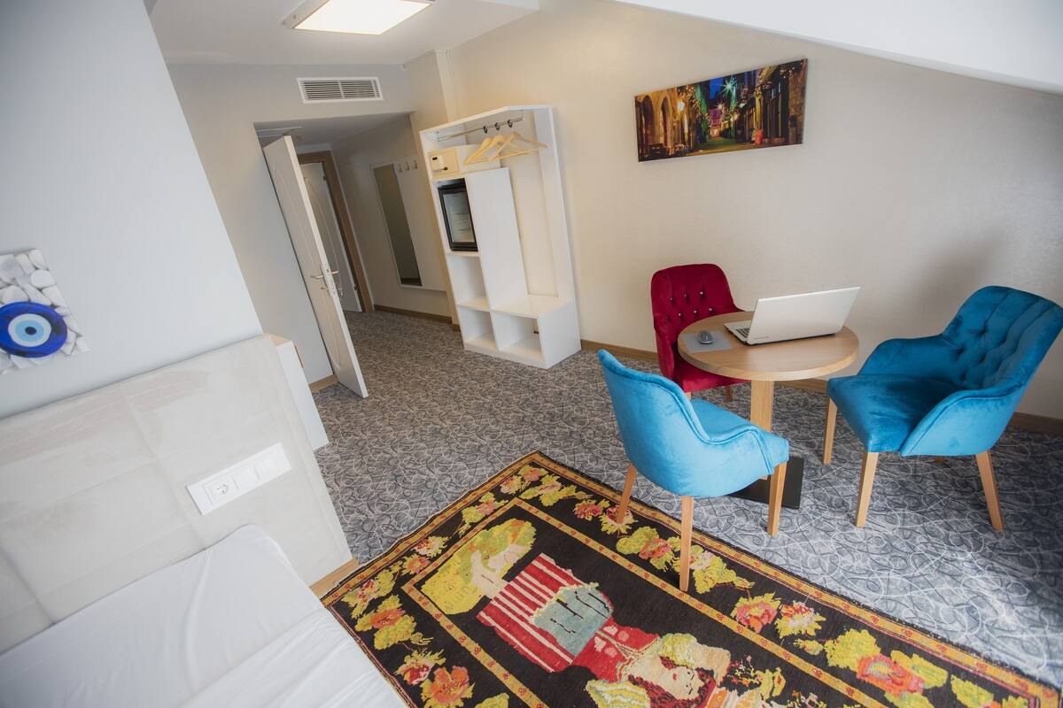 A hotel room with a bed, a desk with a laptop, two blue chairs, a red chair, a white wardrobe, a painting on the wall, and a patterned rug.