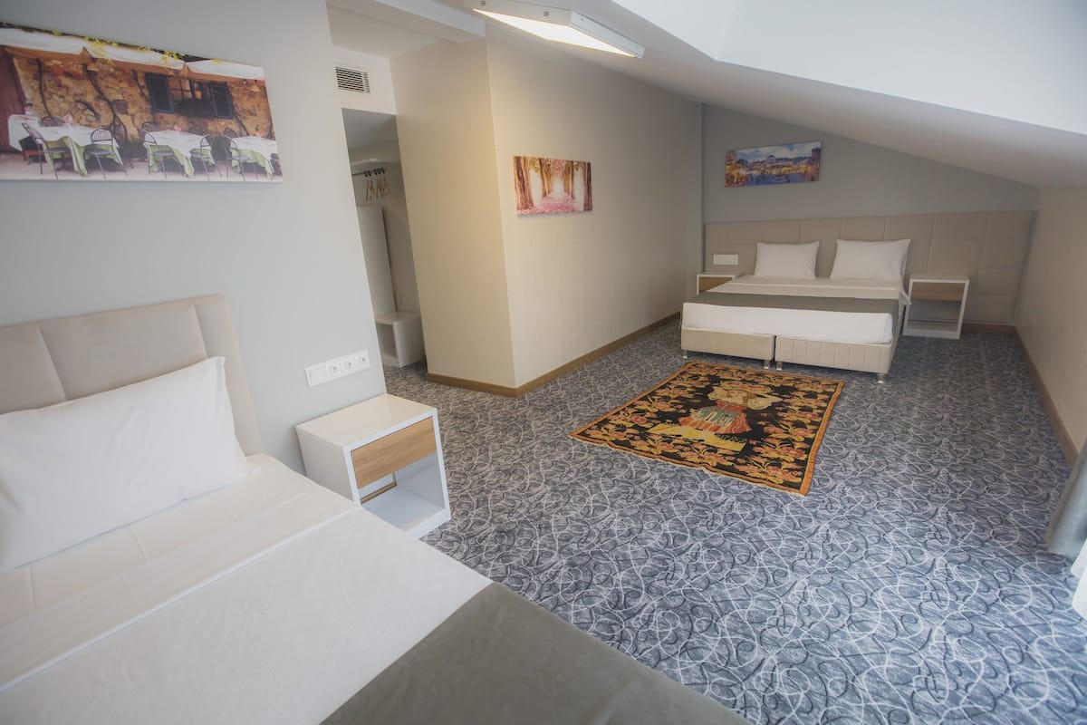 A compact hotel room with a bed, a small bedside table, a nightstand, a decorative rug, and a painting on the wall.