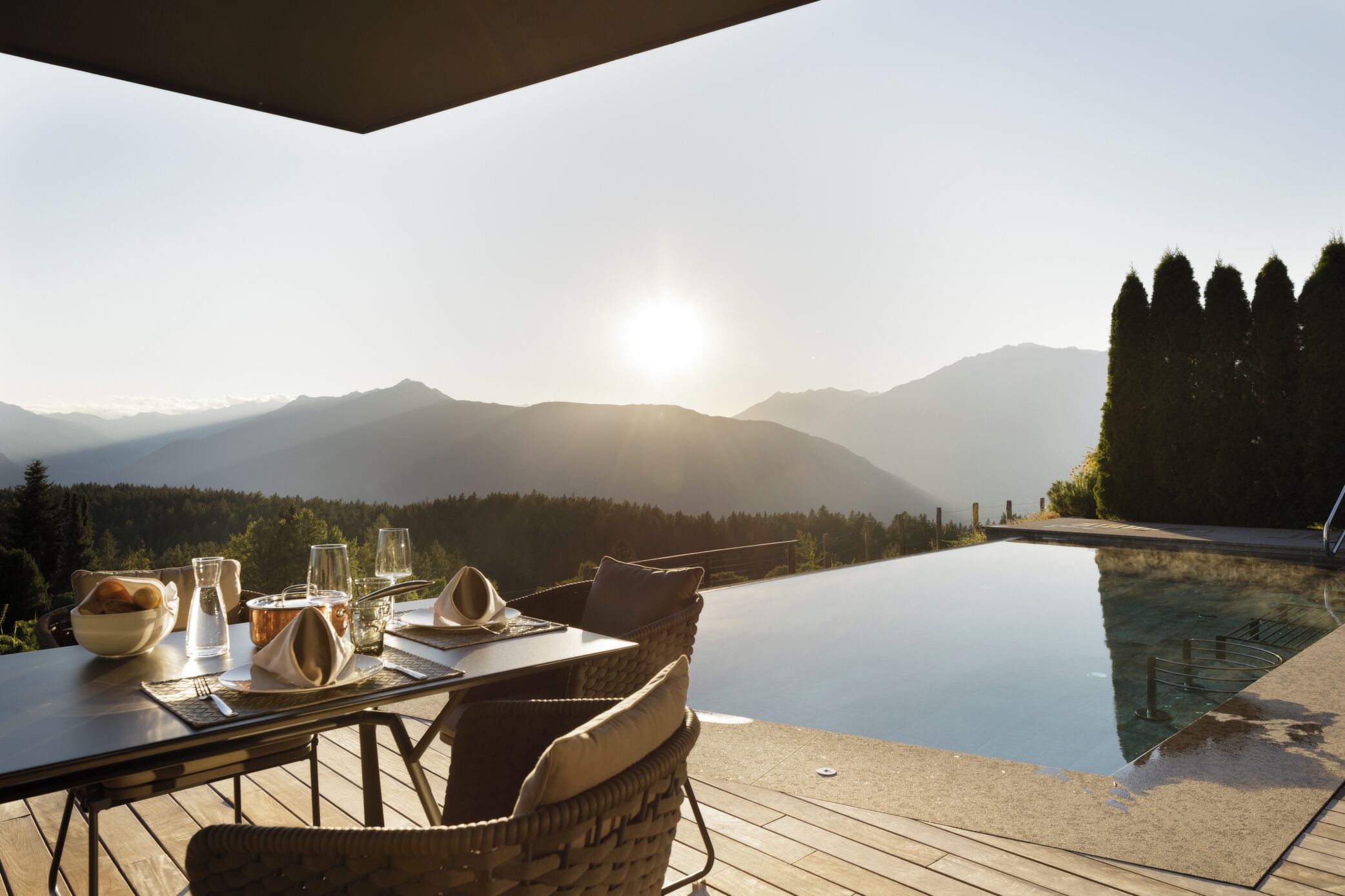 An infinity pool with a mountain view, a dining table set with glasses and a bowl of fruit, and a wicker chair.