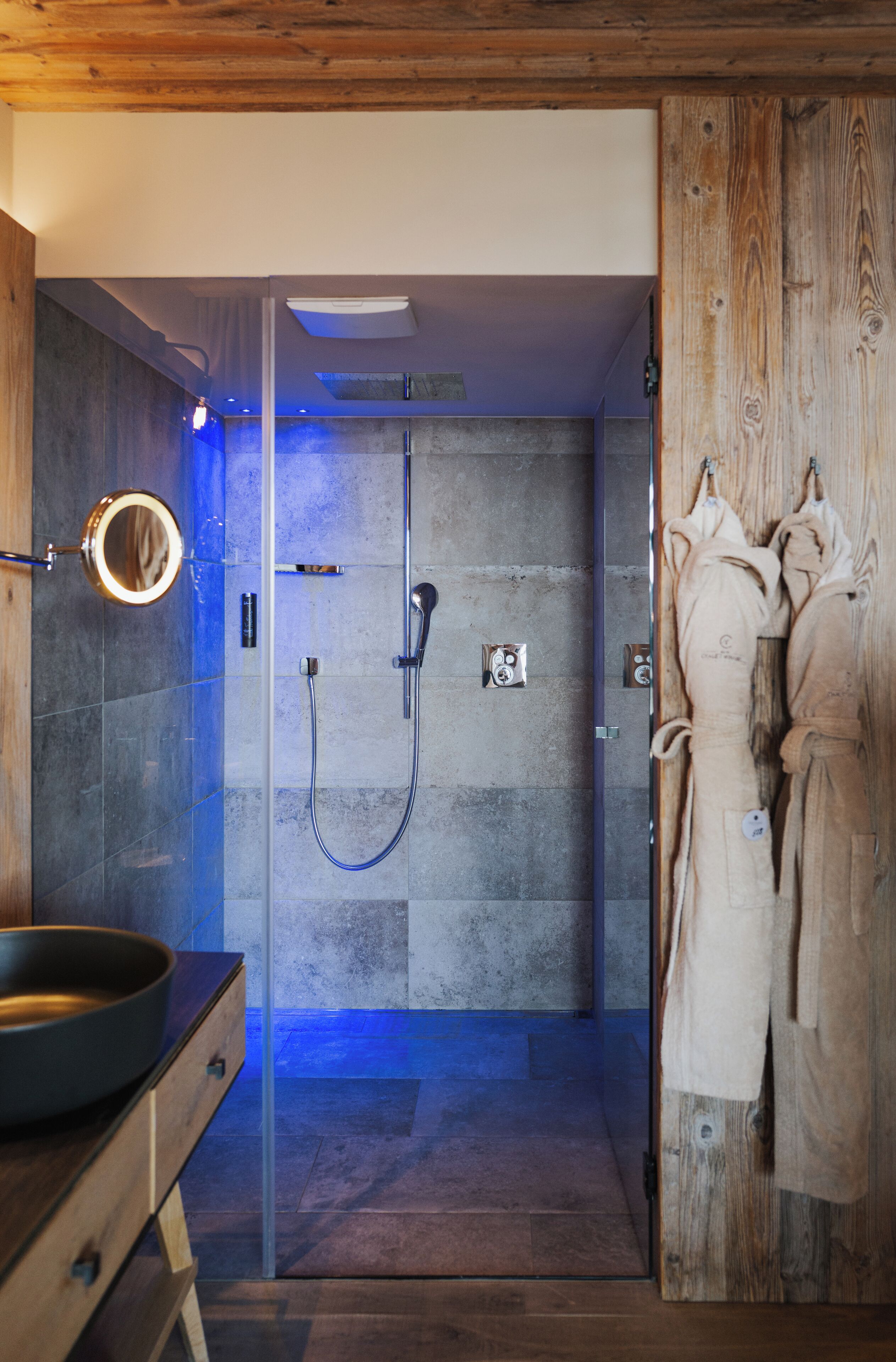 A modern bathroom with a glass shower enclosure, a wooden vanity, and a robe hanging on the wall.