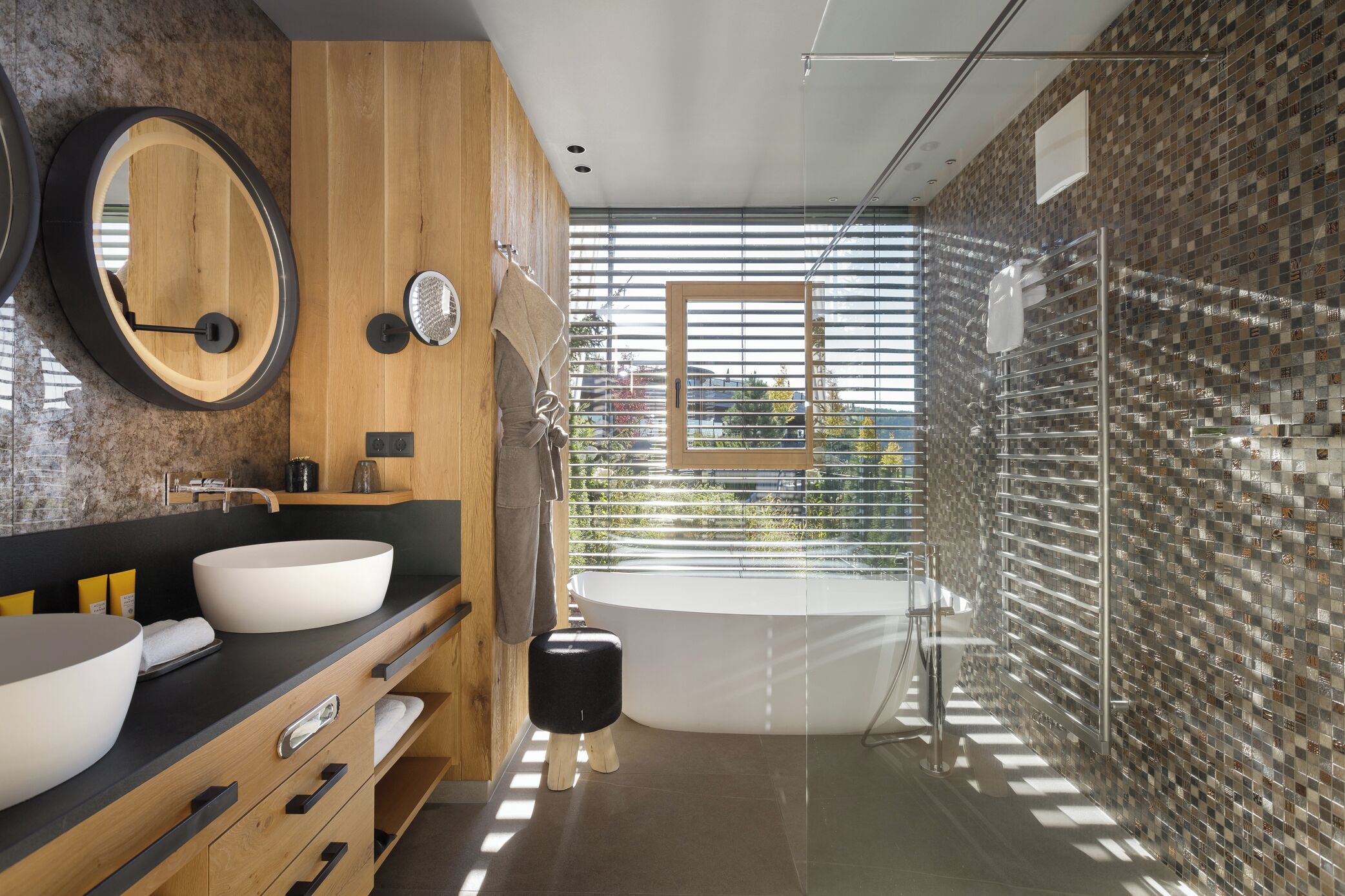 Un bagno moderno con due ampi lavabi, uno specchio rotondo, uno sgabello indipendente, una vasca da bagno e una doccia con pareti in vetro.