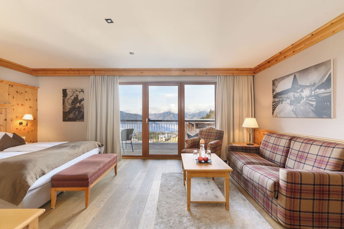 Ett hotellrum med en säng, en soffa, ett bord och utsikt över bergen genom glasdörren.