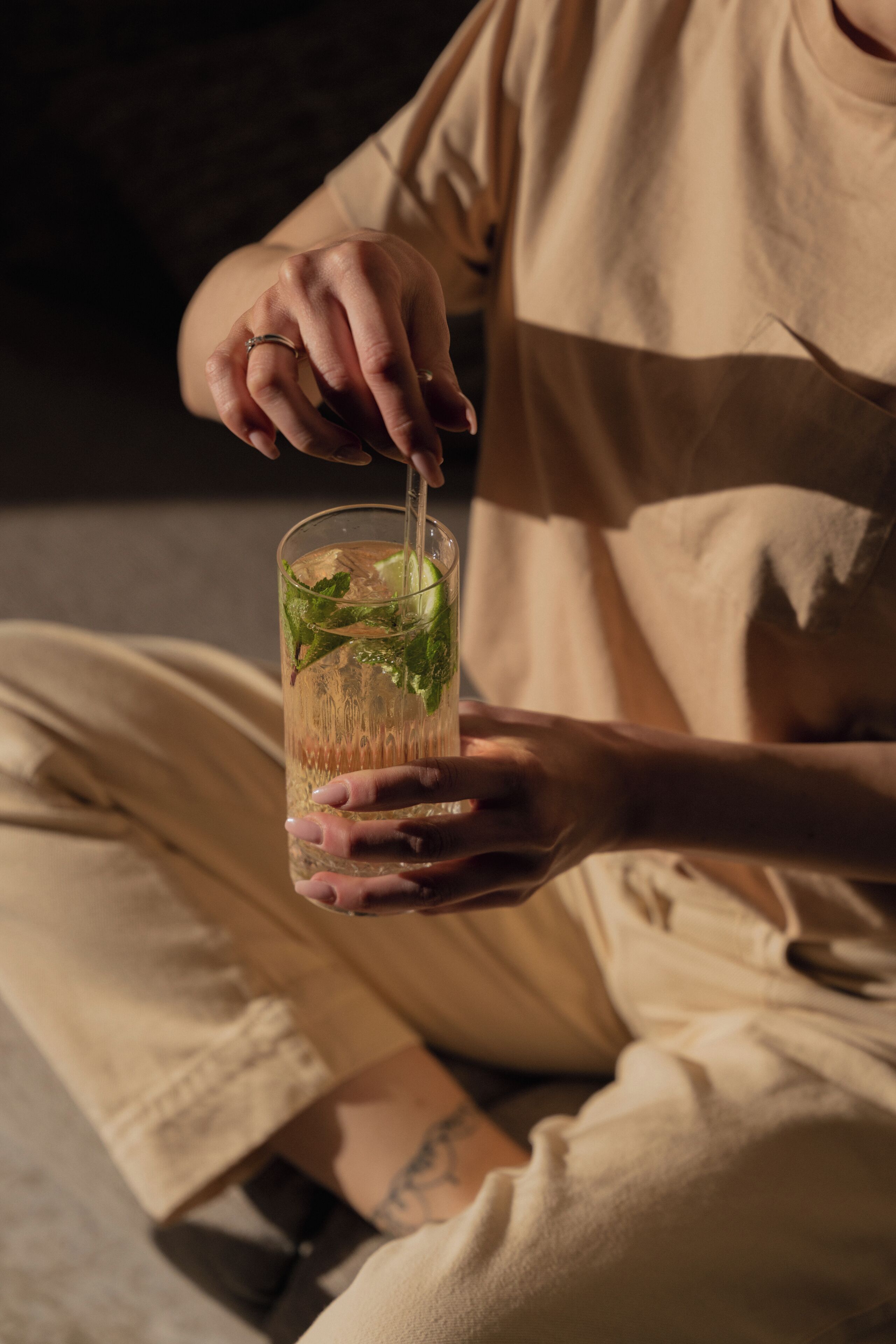 A person holding a glass with a straw, wearing a beige shirt and pants, sitting on a chair with a visible tattoo on their arm.