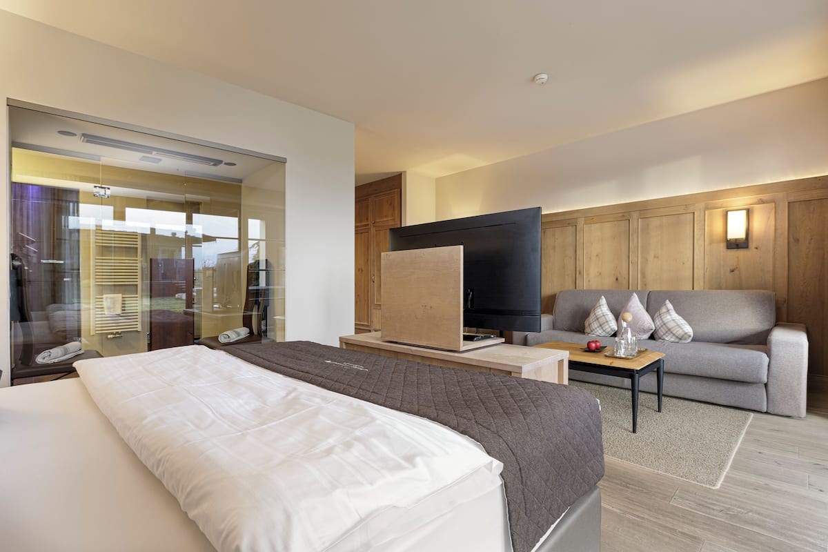 Ett modernt hotellrum med en stor säng, en soffa, en TV och ett badrum med glasväggar.