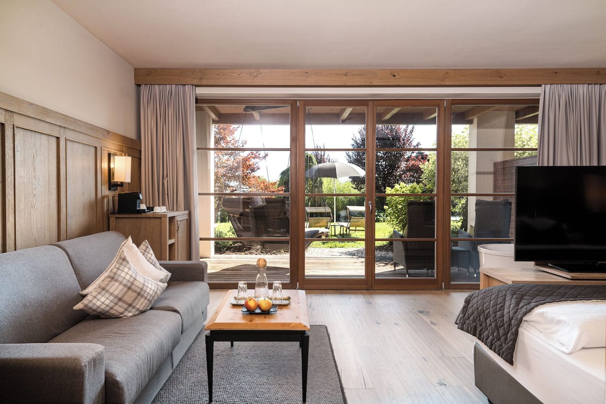 Ett modernt hotellrum med en soffa, ett soffbord, en TV och en skjutdörr i glas som leder ut till ett utomhusområde med en trädgård och en grillplats.