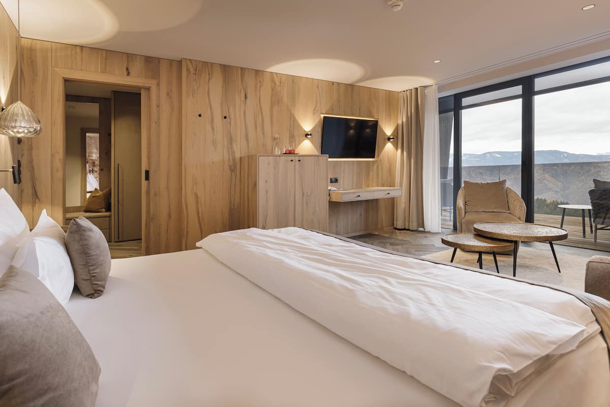 Ett hotellrum med en stor säng, en tv som är monterad på väggen, en väggpanel i trä, en balkong med en stol och ett bord samt utsikt över bergen.