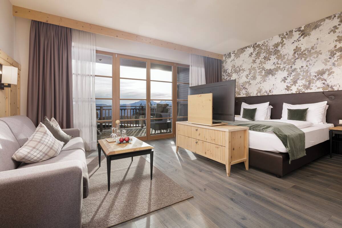Ett modernt hotellrum med en soffa, ett träbord vid sängen, en säng med tv och en balkong med utsikt.