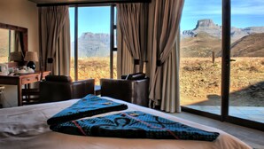 A hotel room with a large bed, a desk with a lamp, and a view of a mountainous landscape through large windows. - Witsieshoek Mountain Lodge (Maluti a Phofung)