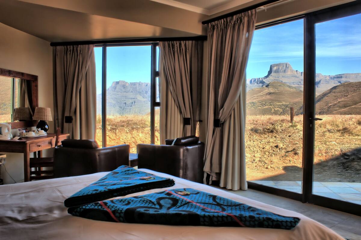 A hotel room with a large bed, a desk with a lamp, and a view of a mountainous landscape through large windows.