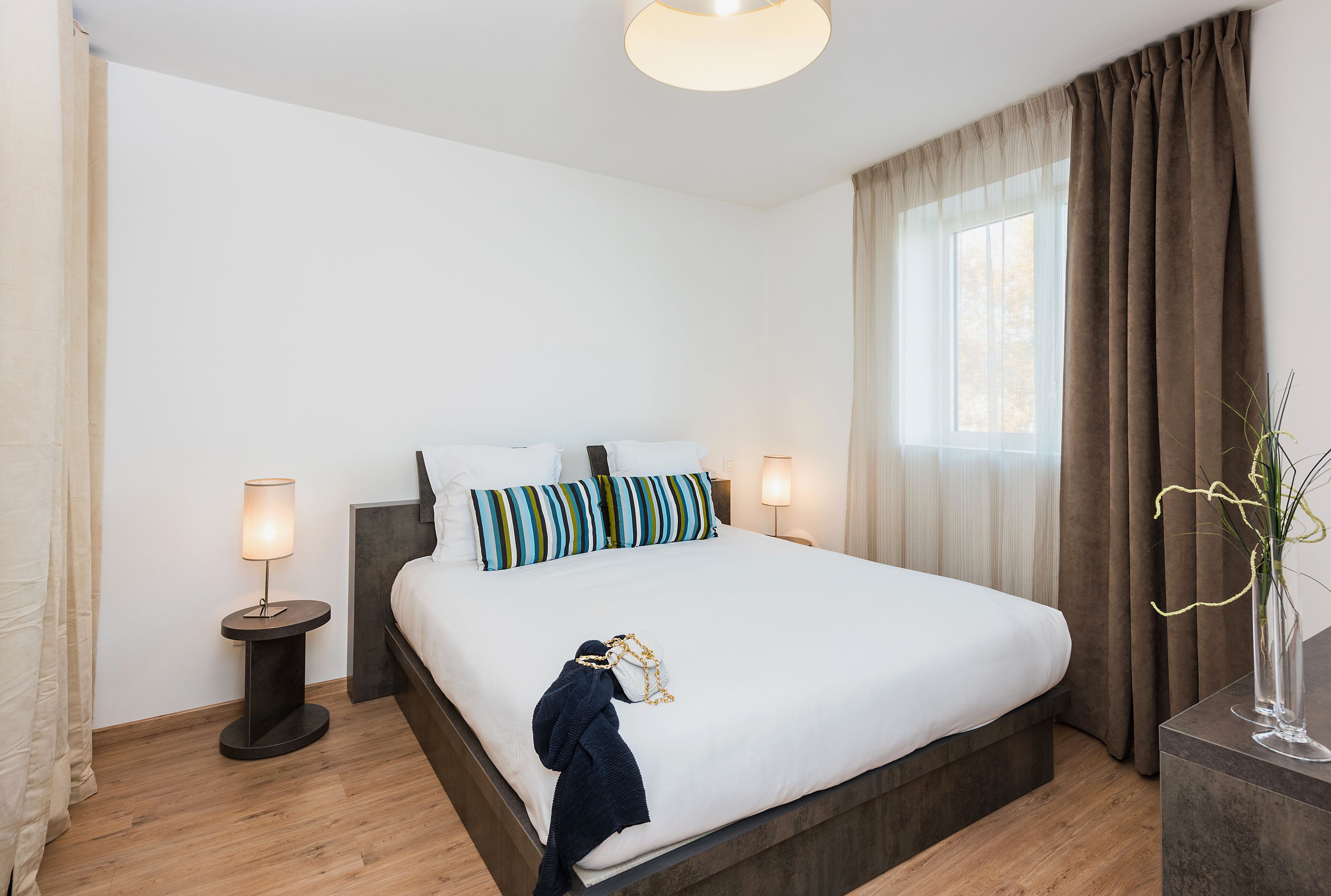 A neatly made bed with striped pillows, a bedside lamp, and a vase with plants on a nightstand.