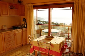 A dining area with a table set for two, a view of the sea, and a kitchen with wooden cabinets.