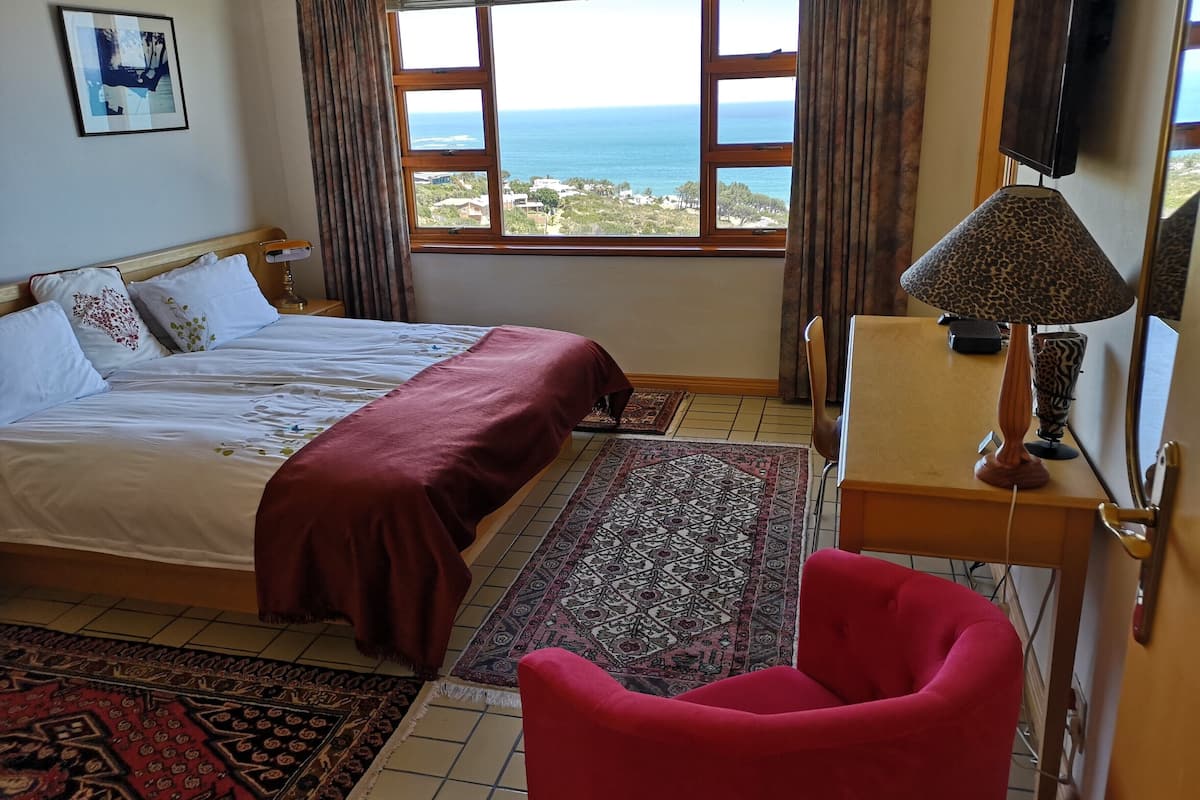 A bedroom with a bed, a desk, a chair, a TV, and a view of the sea.