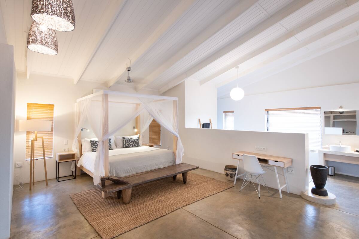 A bedroom with a four-poster bed, a desk, and a bathroom with a mirror and sink.