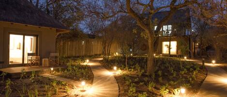 A well-lit garden pathway leading to a building with a thatched roof and a large glass door.