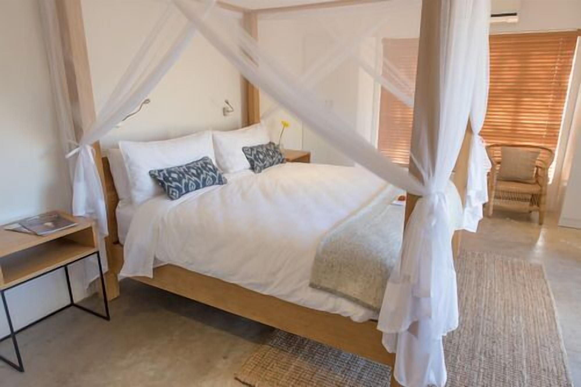 A bedroom with a canopy bed, a bedside table, a chair, and a window with blinds.