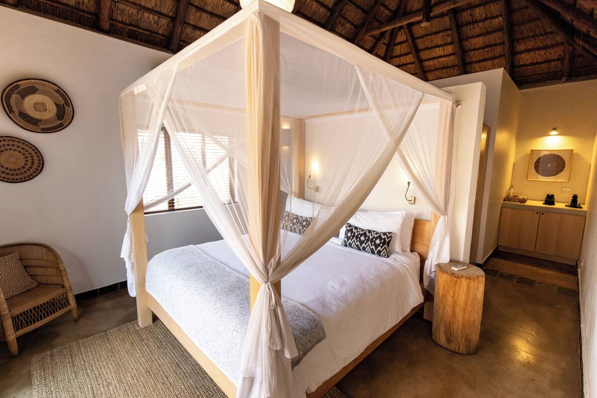 A four-poster bed with a canopy, a wicker chair, a wooden stool, and a kitchen area with cabinets and a countertop.