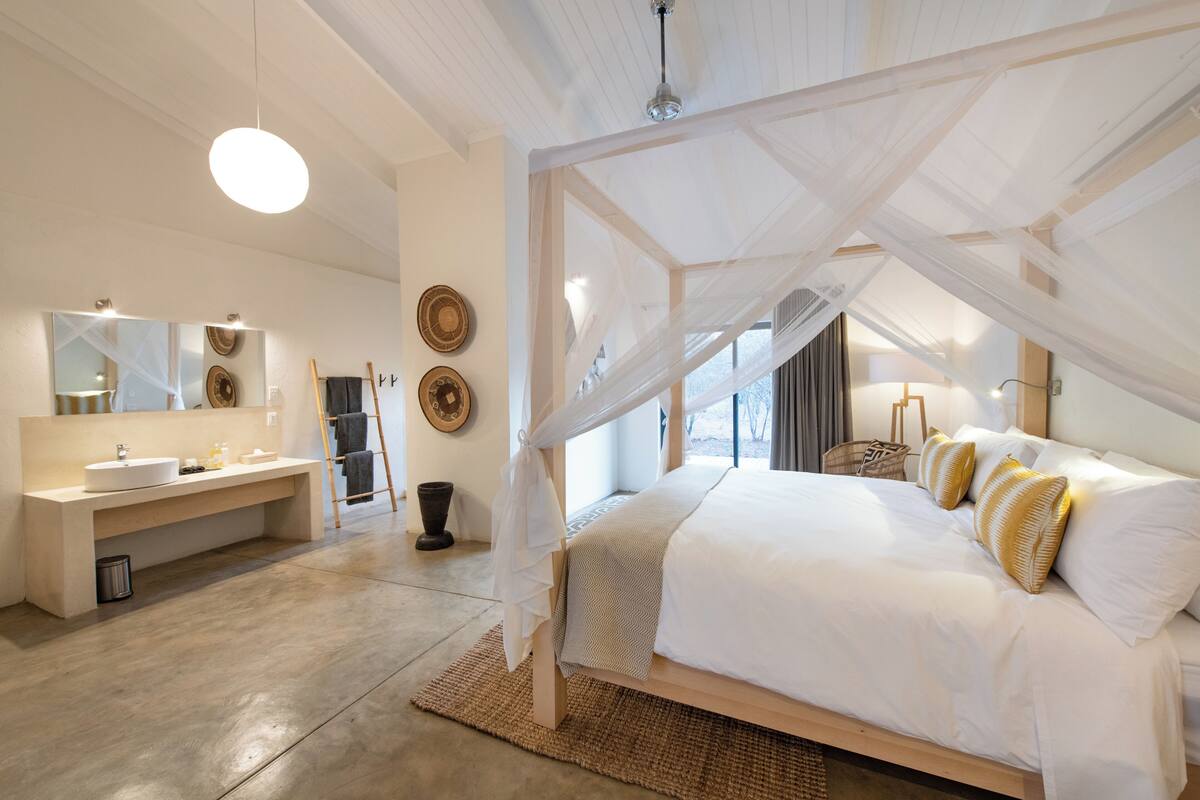 A bedroom with a canopy bed, a vanity with a mirror, and a large window.