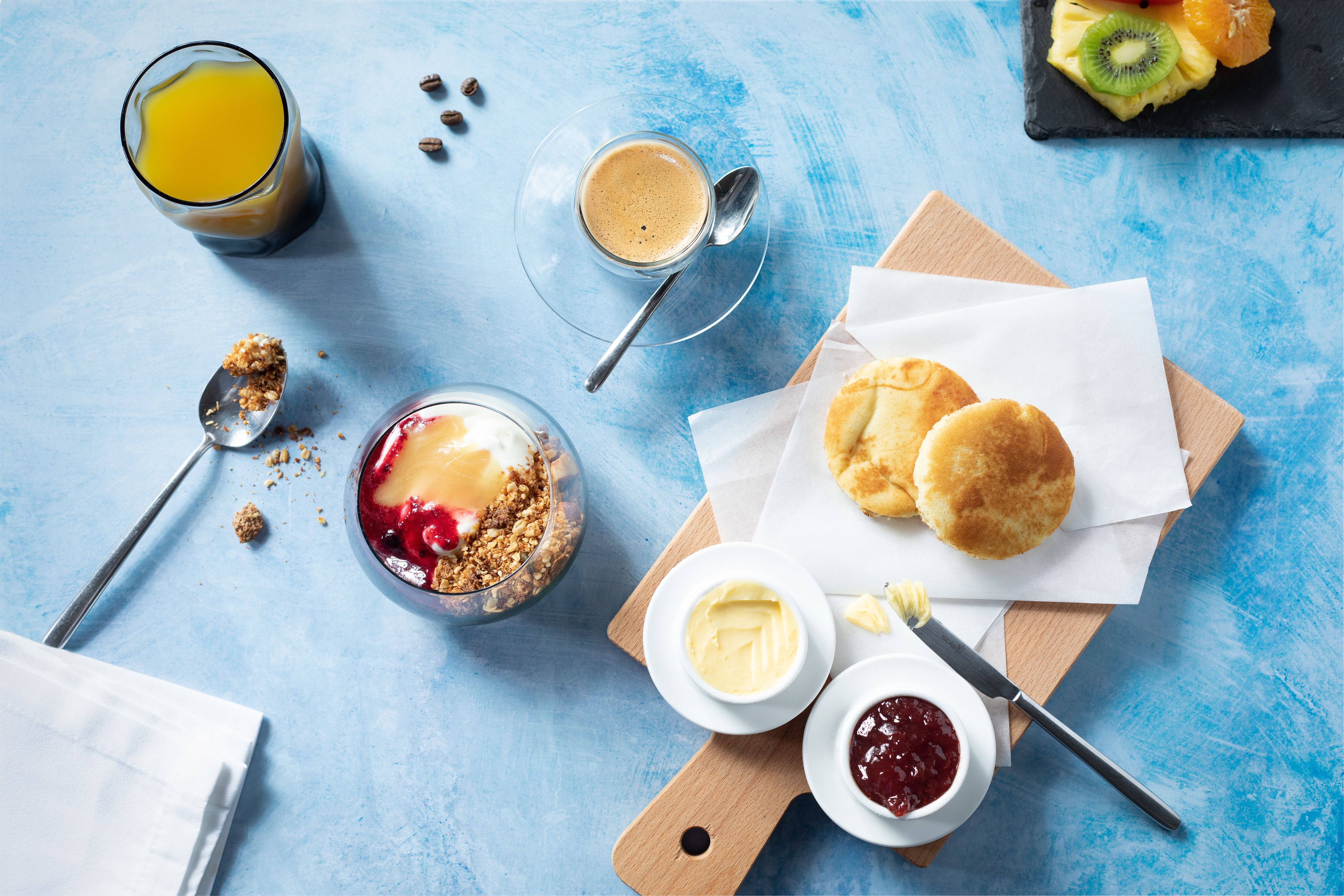 A breakfast setting with a glass of orange juice, a bowl of cereal with fruit, two biscuits, and condiments on a wooden board.